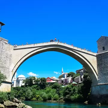 A single high stone arch spanning a green river between two fortified towers, onlookers along the parapet and a minaret in the old town beyond.