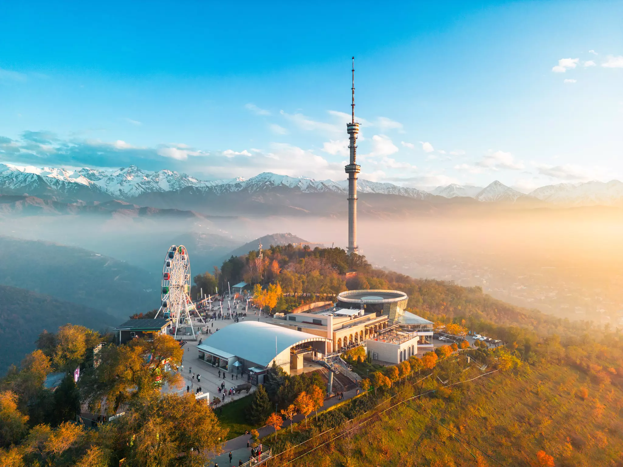An aerial view of an amusement park and television tower at the top of a hill, shrouded in mist. Snow-capped mountains are seen in the distance.