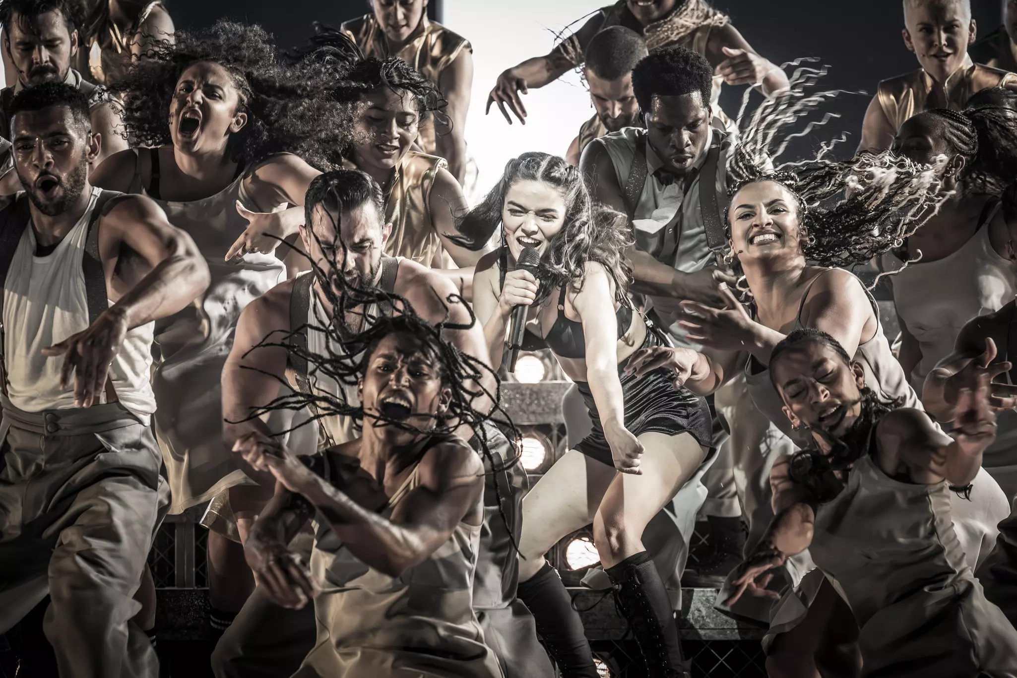 A woman with a microphone sings as she is surrounded by a group of dancers in an energetic stage performance