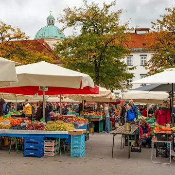 Shoppers browse produce under white and red umbrellas, with a green church dome and autumn trees behind