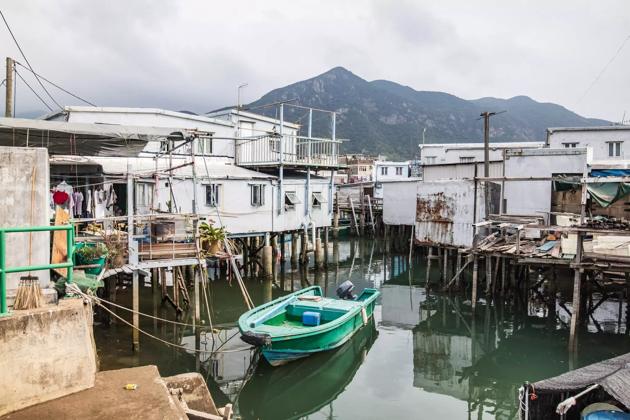 A green boat is moored in a small channel surrounded by houses on stilts in the water.