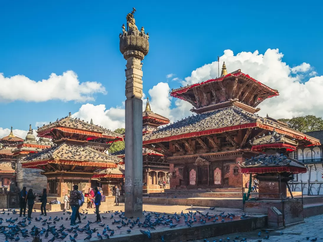 Brick temples with red-trimmed tiered roofs and a tall stone pillar topped by a statue, pigeons and people below.