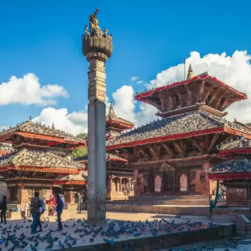 Brick temples with red-trimmed tiered roofs and a tall stone pillar topped by a statue, pigeons and people below.