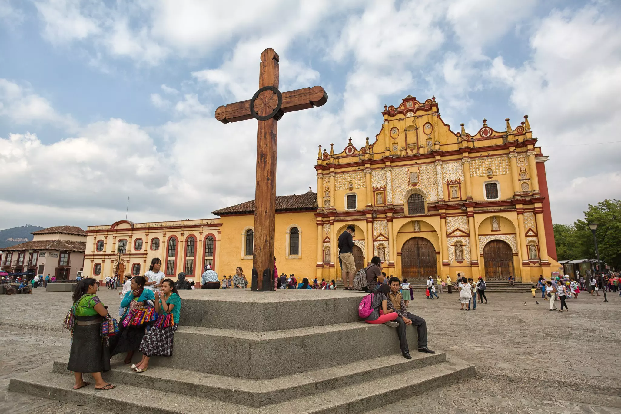 People congregate in Plaza de la Paz in front of San Cristóbal cathedral, a yellow building with red trim. There is a large wooden cross in the foreground.