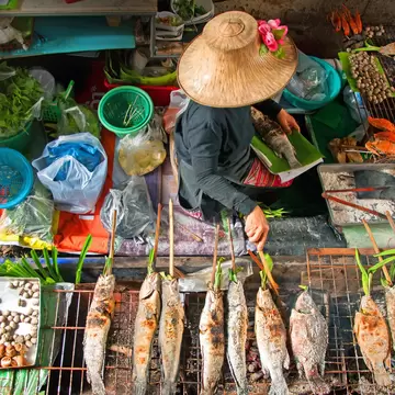 Vendor in a flower-trimmed straw hat grilling skewered fish in a boat, with trays of cockles, snails, prawns and crab