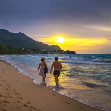 Two people walking at the water's edge on golden sand as the sun sets beside a forested Mahe headland