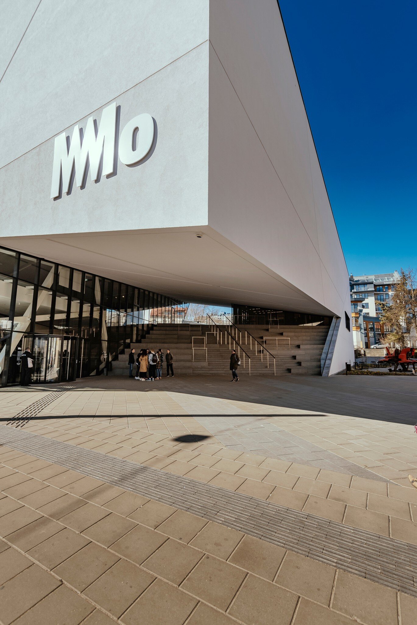 Large white cantilevered building with a MO logo over a glass entrance, people gathered by wide stone steps