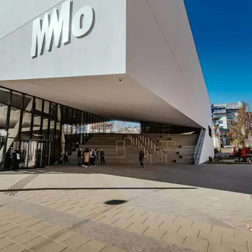 Large white cantilevered building with a MO logo over a glass entrance, people gathered by wide stone steps