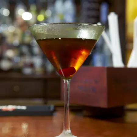 Close-up of a triangular-shaped glass with long stem containing a bourbon cocktail with cherry at the bottom on a wooden table.