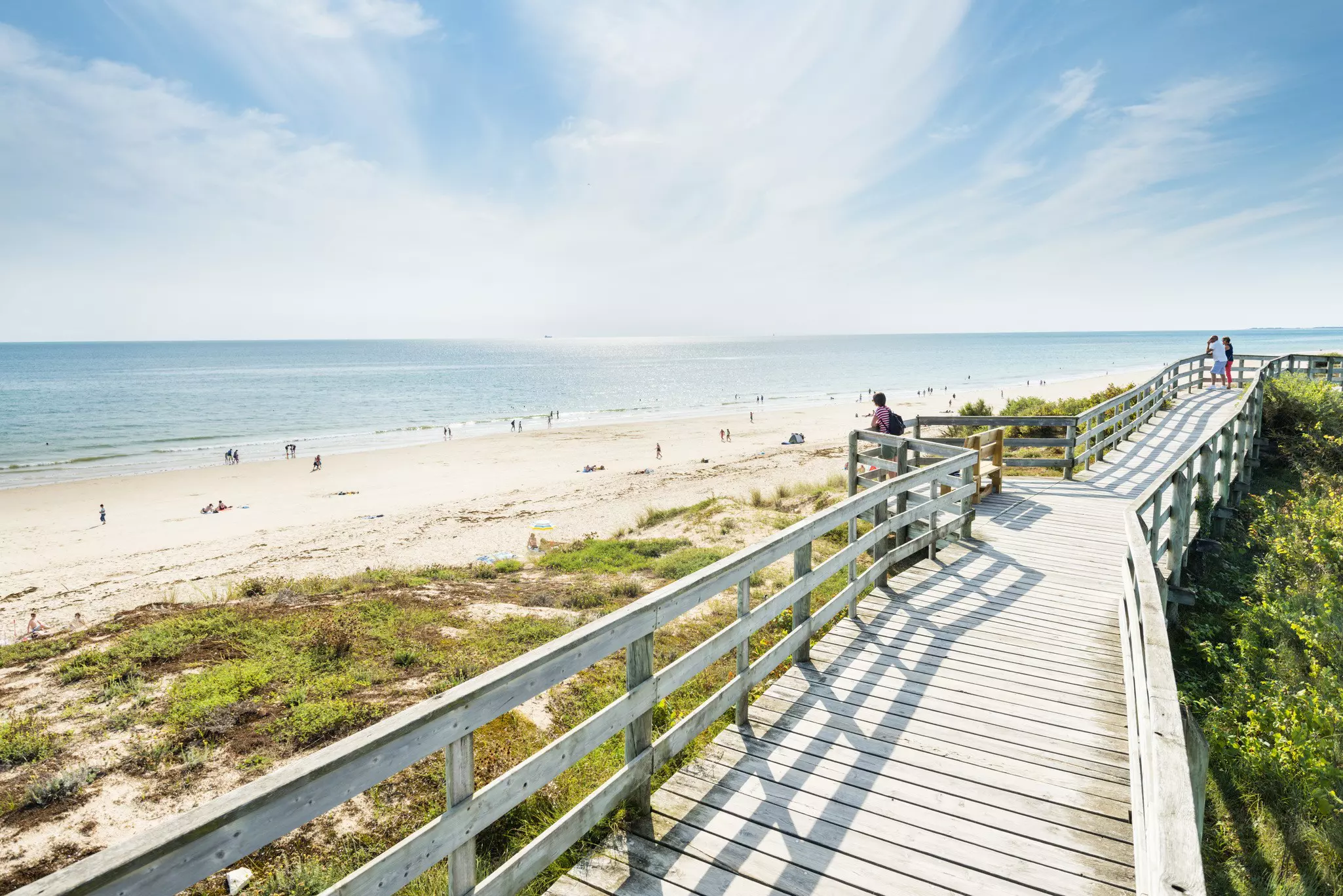 A long boardwalk above a sand dune overlooking a sandy beach where people are relaxing or playing at the edge of the water.