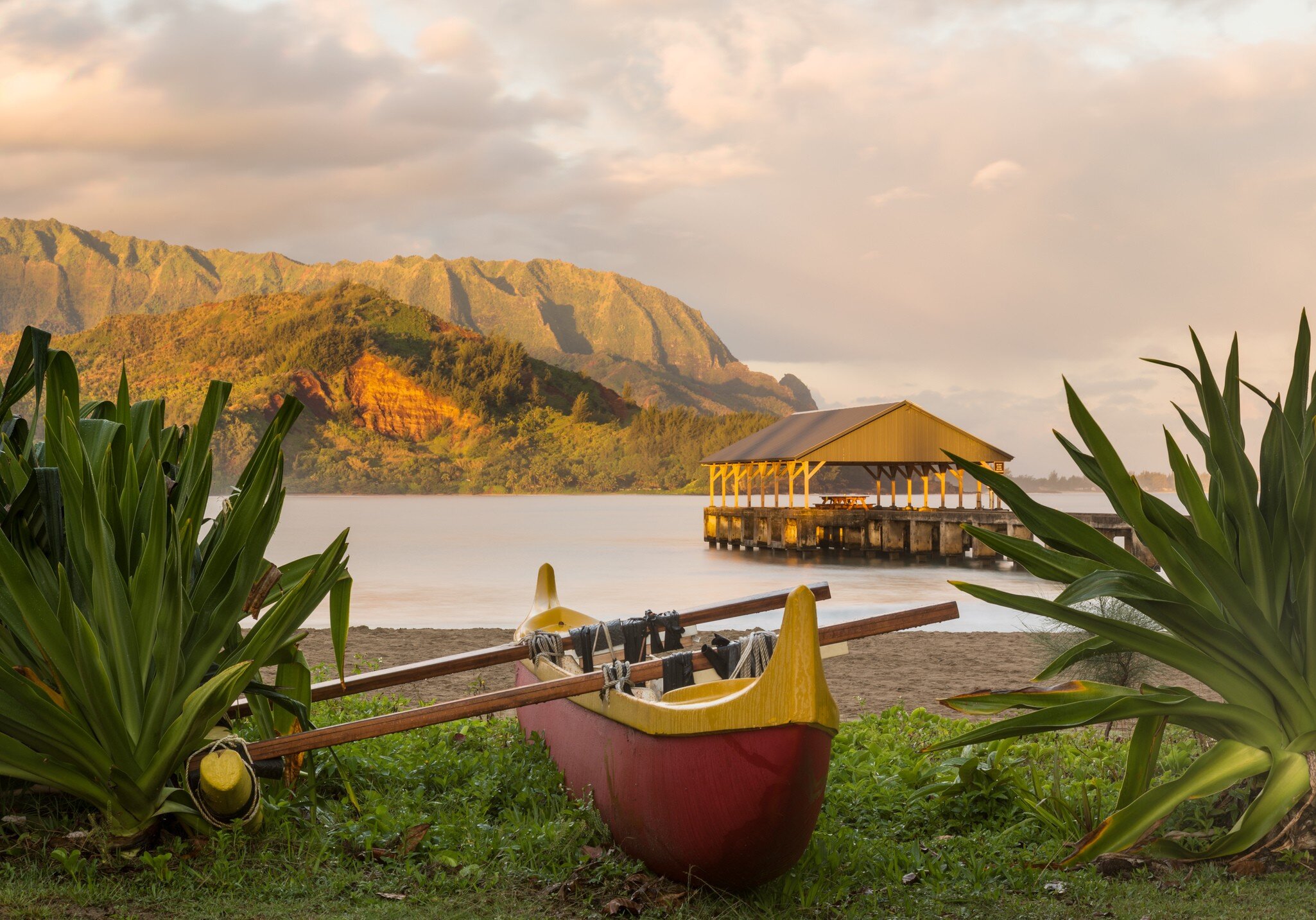 Red and yellow hawaiian canoe with outrigger on the beach at Hanalei pier at dawn as the sun lights the sky over Na Pali mountains