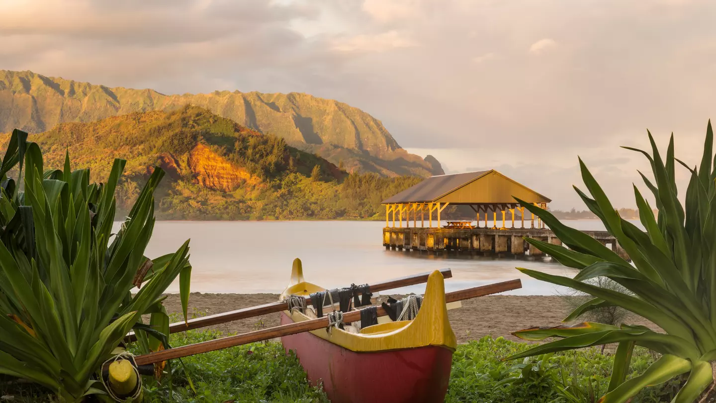 A wooden canoe sits on the edge of a beach with a covered pier, backed by mountains
