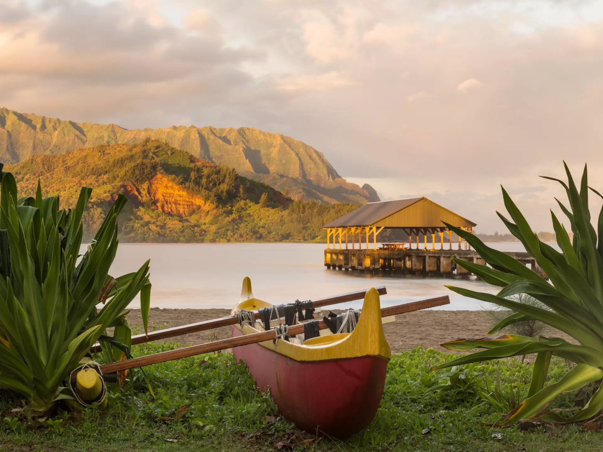 A wooden canoe sits on the edge of a beach with a covered pier, backed by mountains