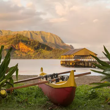 A wooden canoe sits on the edge of a beach with a covered pier, backed by mountains