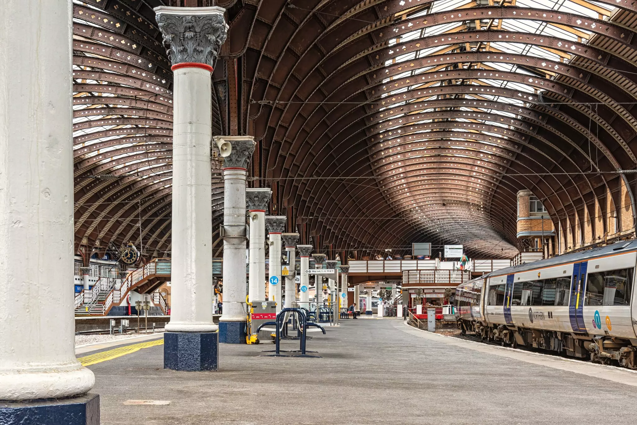A railway station platform leads to a footbridge with a balcony. There are columns supporting the vast curved station roof.