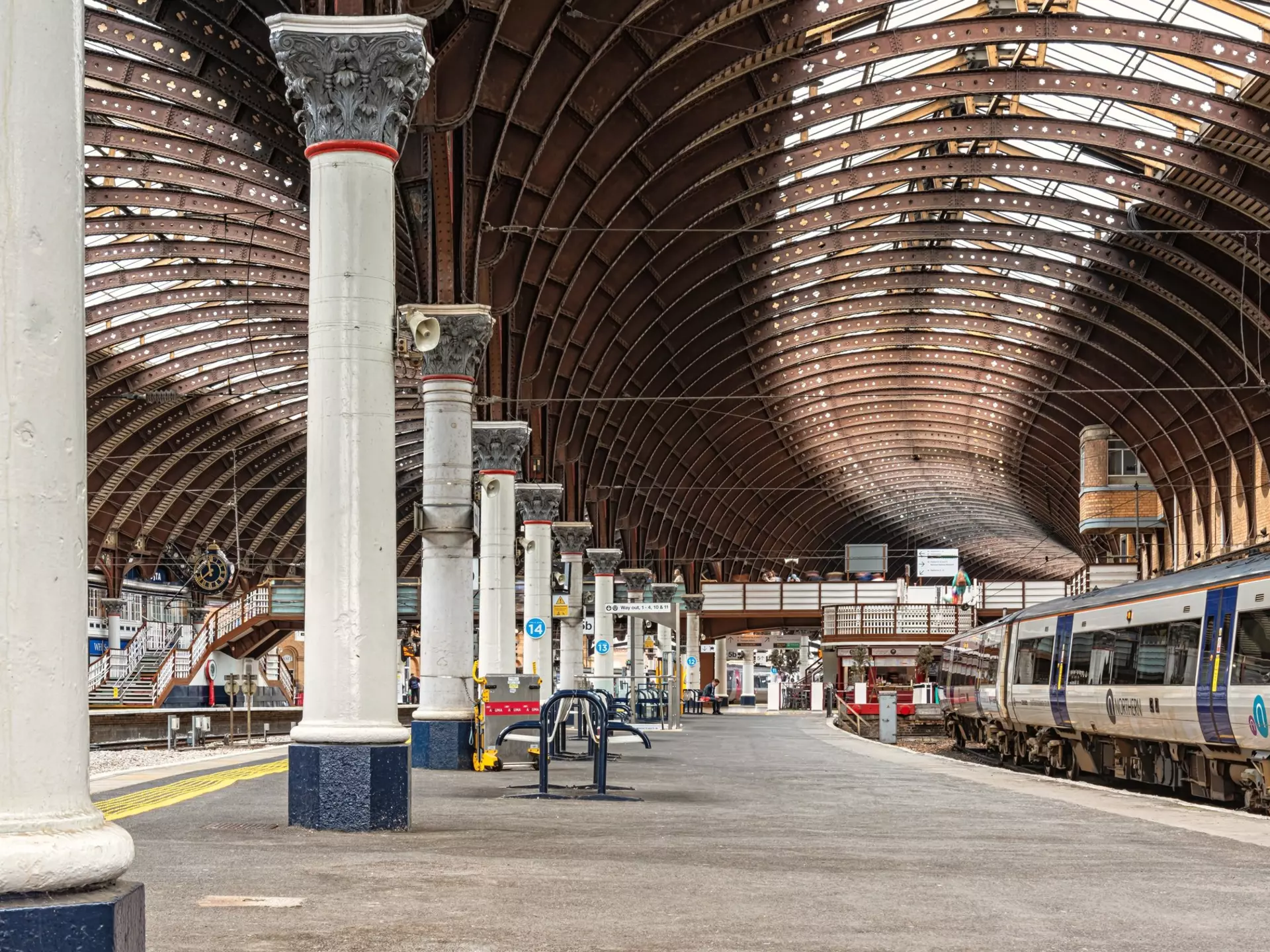 A railway station platform leads to a footbridge with a balcony. There are columns supporting the vast curved station roof.