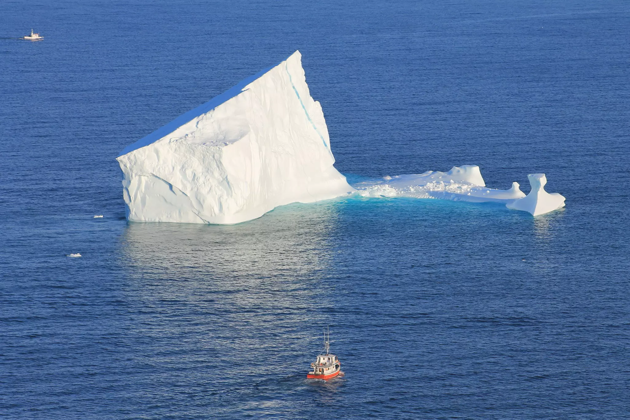 Iceberg floating in the sea with an orangey-red and white boat in the foreground on a sunny day.