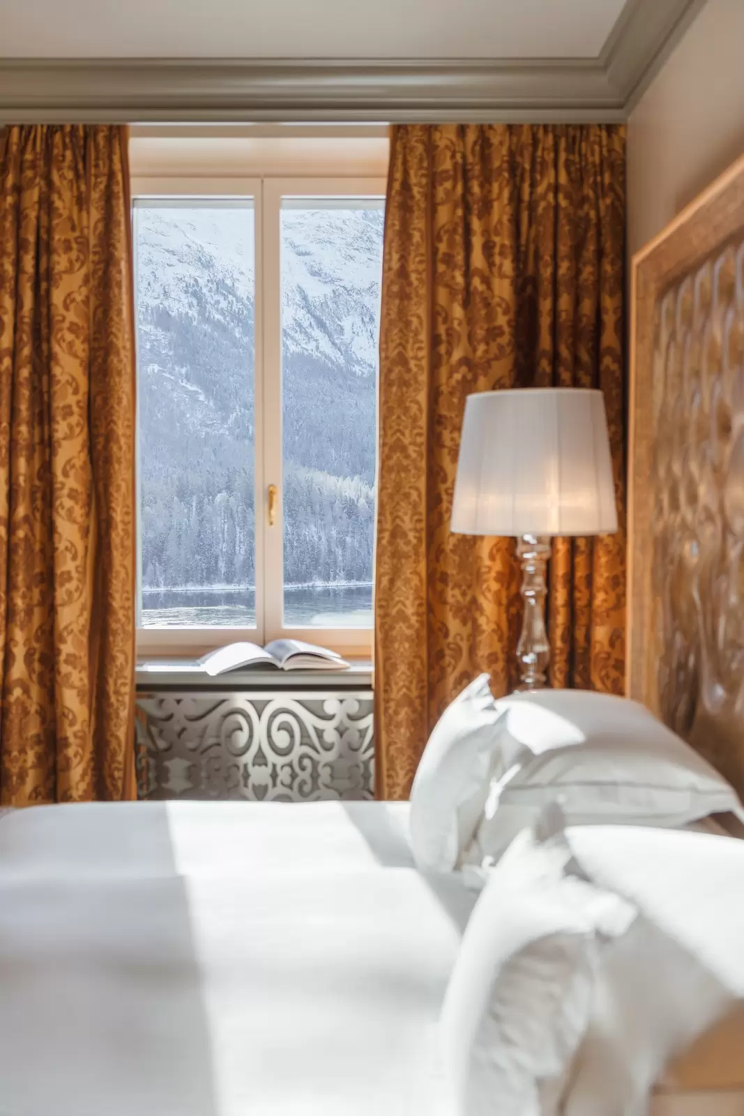 A white bed in a hotel with heavy golden curtains; a snowy landscape is on the other side of the window.