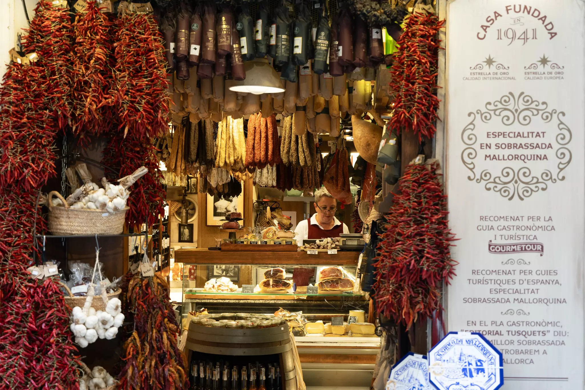 Dried red peppers and garlic hang in the doorway to a shop where a shopkeeper is behind a glass display case, above which hang sausages.