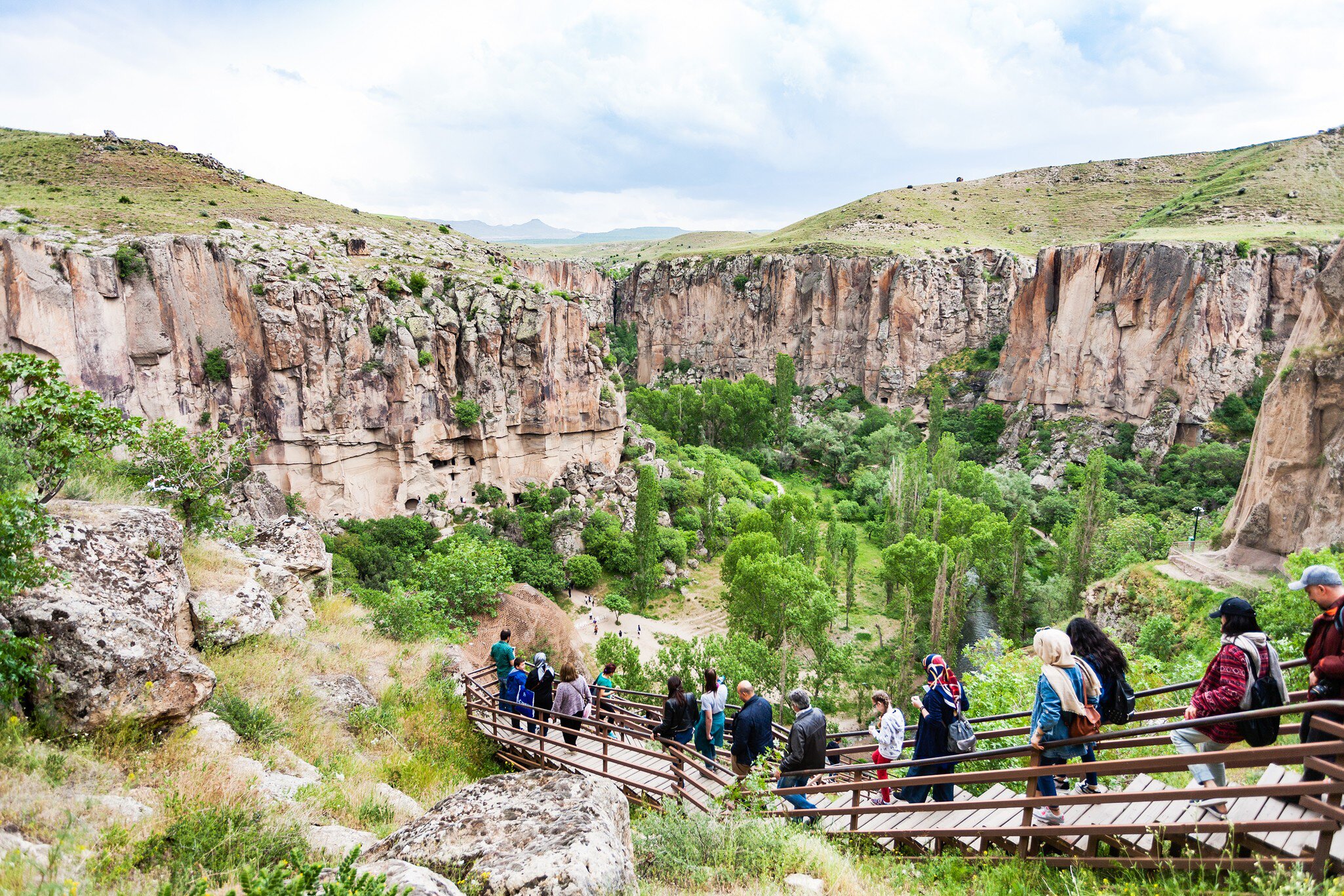 Visitors descending a wooden stairway into a green river gorge walled by banded rock cliffs.