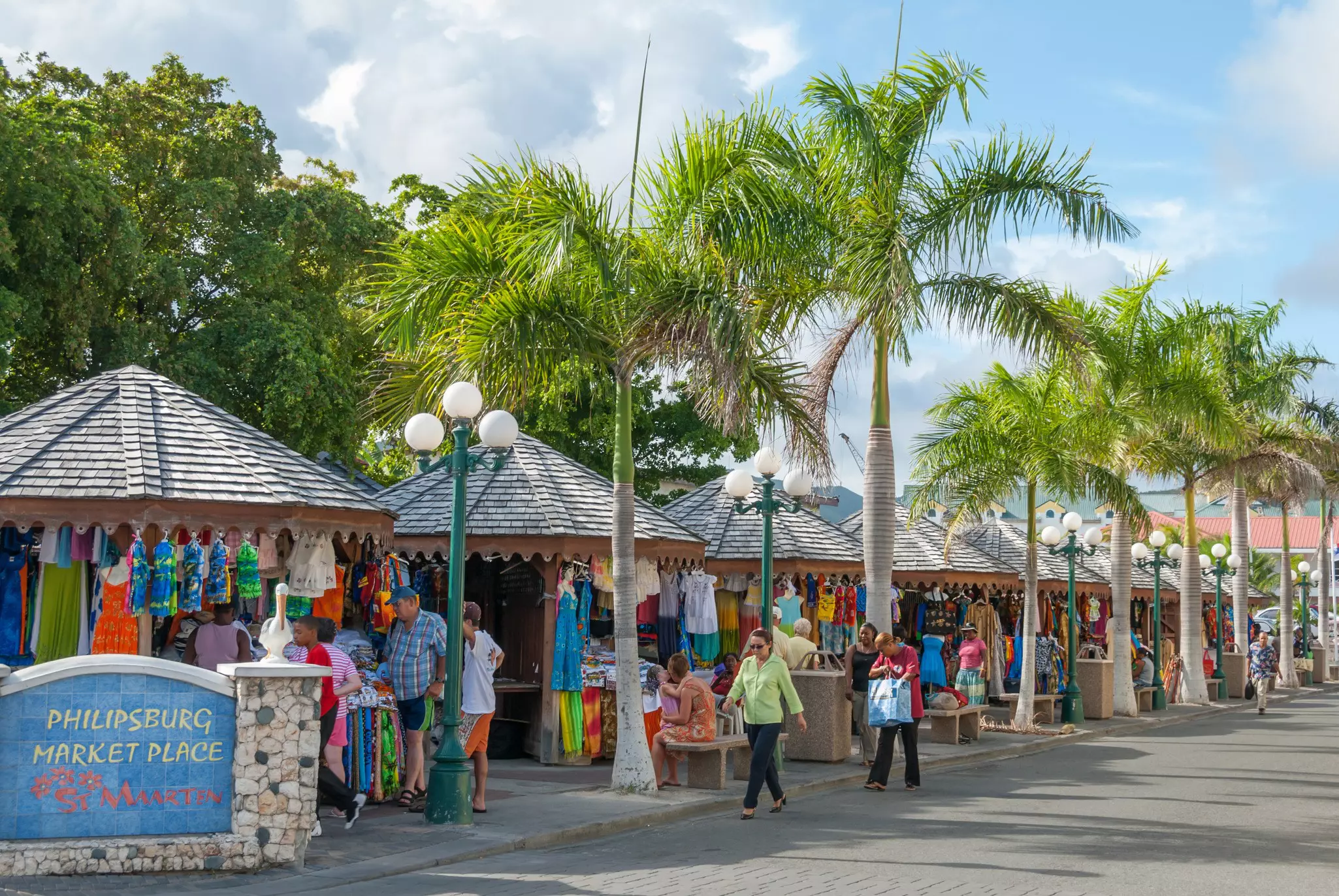 People walking around and browsing at covered kiosks at an outdoor market. Palm trees line the walkway in front of the kiosks