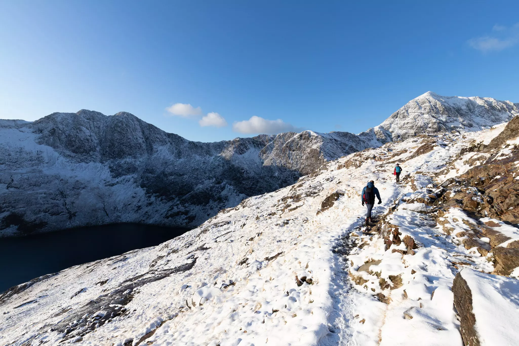 Two people walking along a narrow snow-covered path in the mountains on a sunny day.