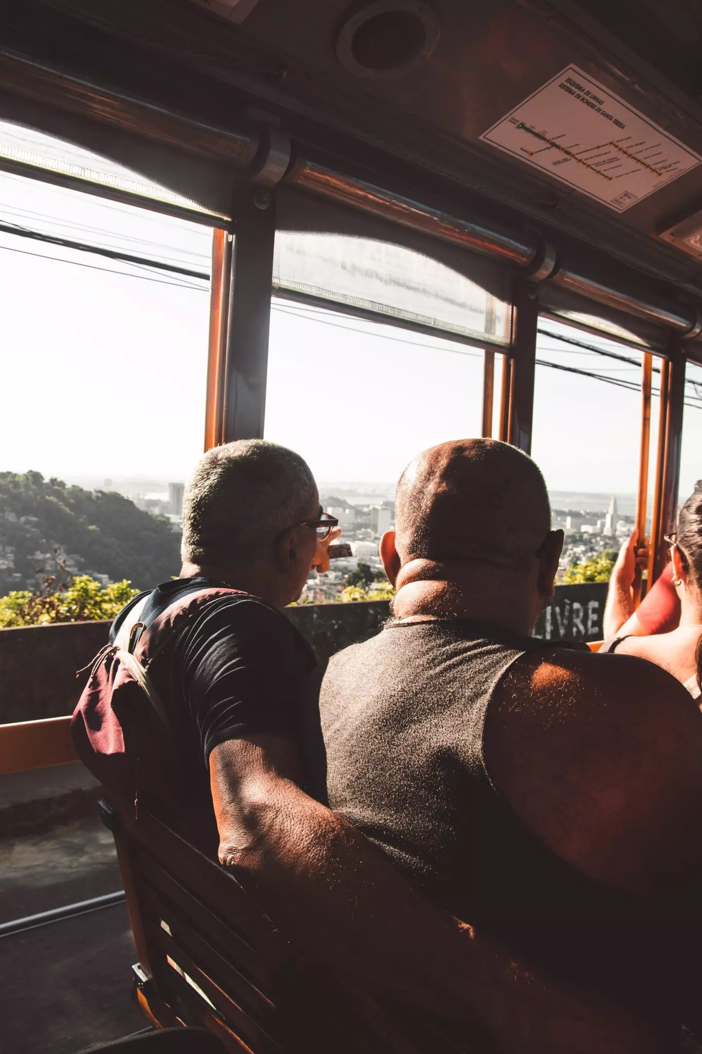 Two people are seen from behind in a tram. A few of hills and buildings in a city is visible through the tram’s windows.