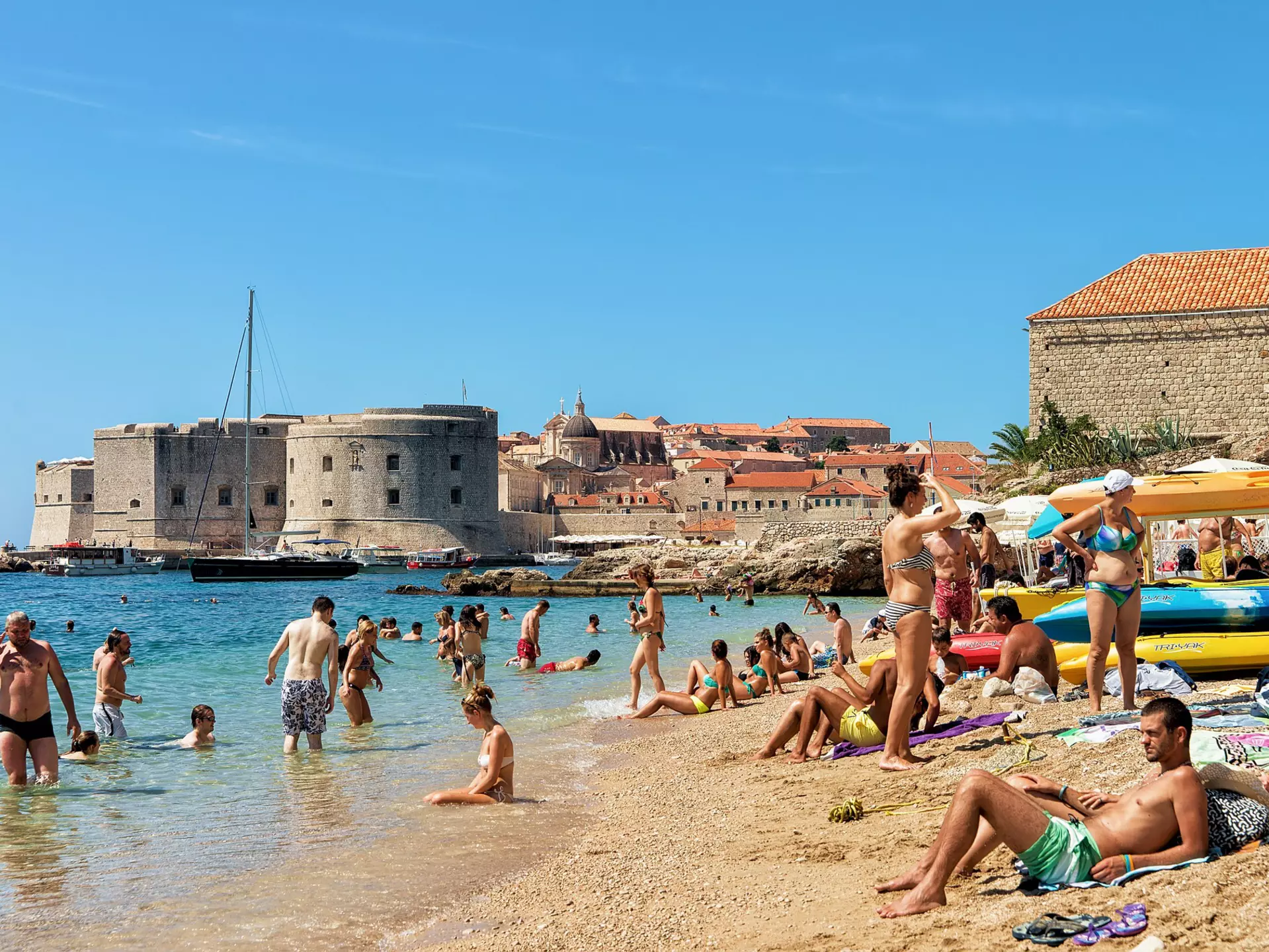 People lie on the sand and wade into the water at a beach by a historic town. A fortress is visible across the water.