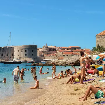 People lie on the sand and wade into the water at a beach by a historic town. A fortress is visible across the water.