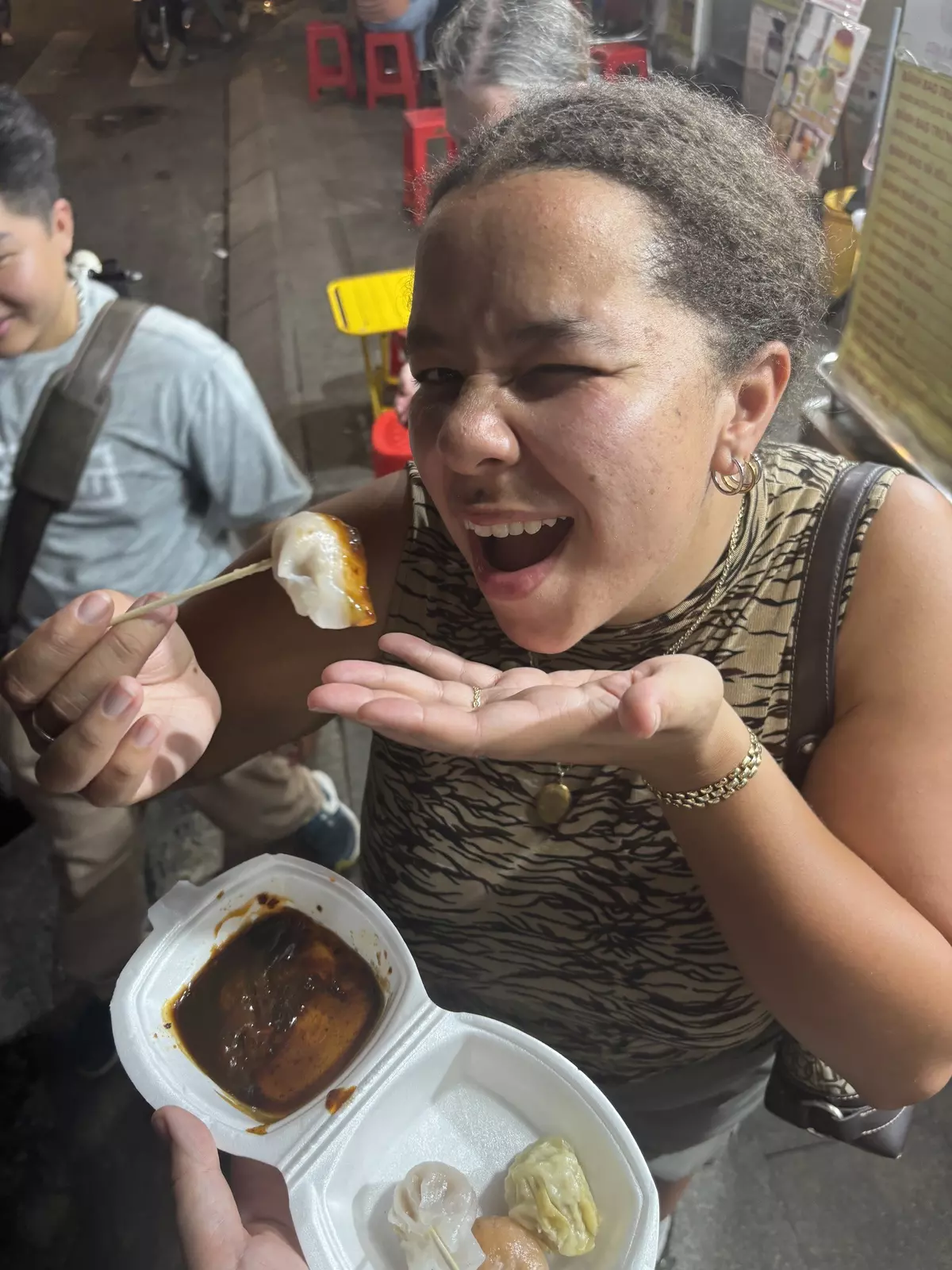 A woman eating a dumpling from a styrofoam container that has sauce and other dumplings in it