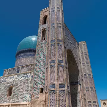 A towering blue-patterned brick portal with a deep arch rises beside a turquoise dome under a deep blue sky.