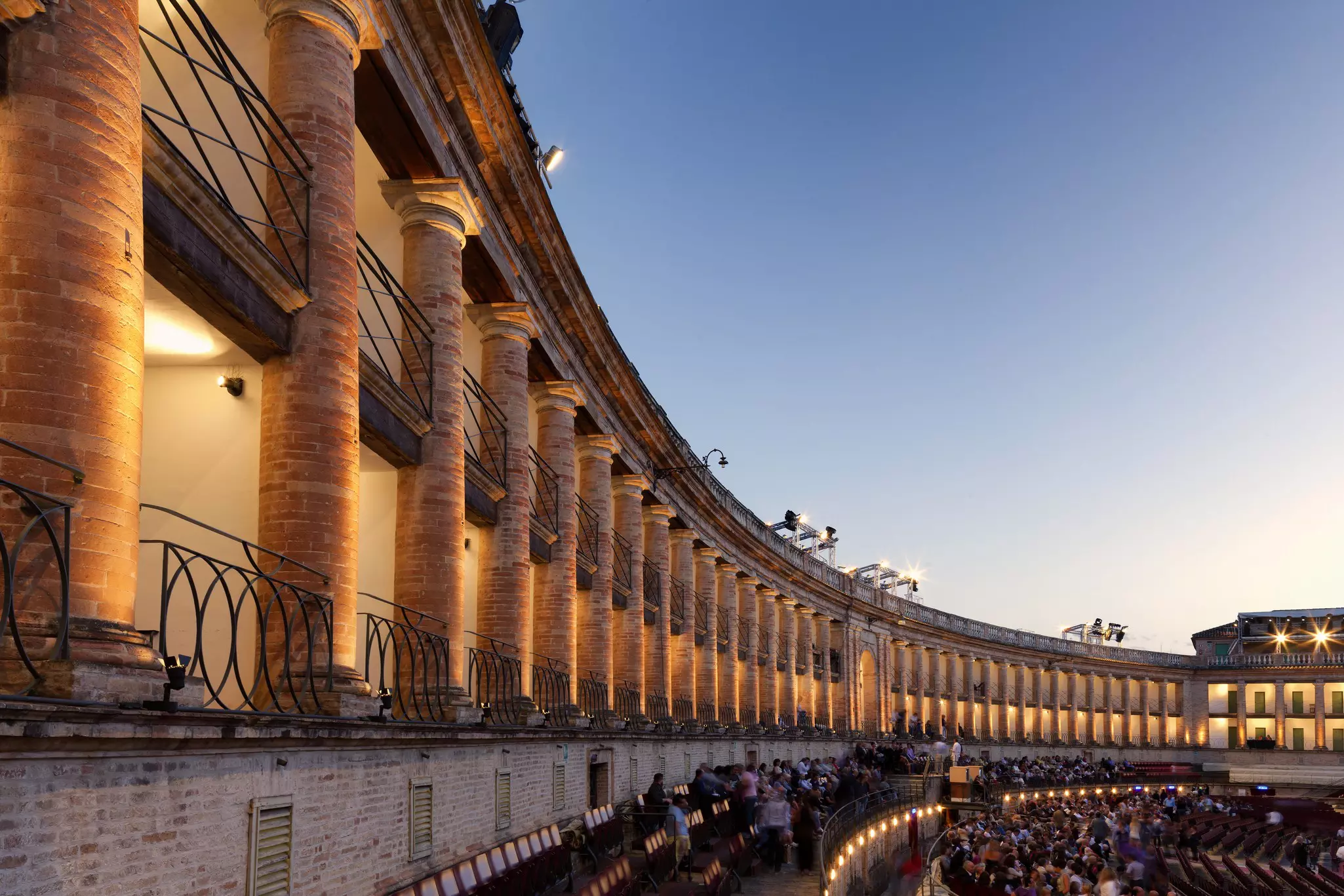 Dozens of columns surround a large outdoor amphitheater; people are seated in front of the wall and the columns are uplit. Le Marche, Italy.