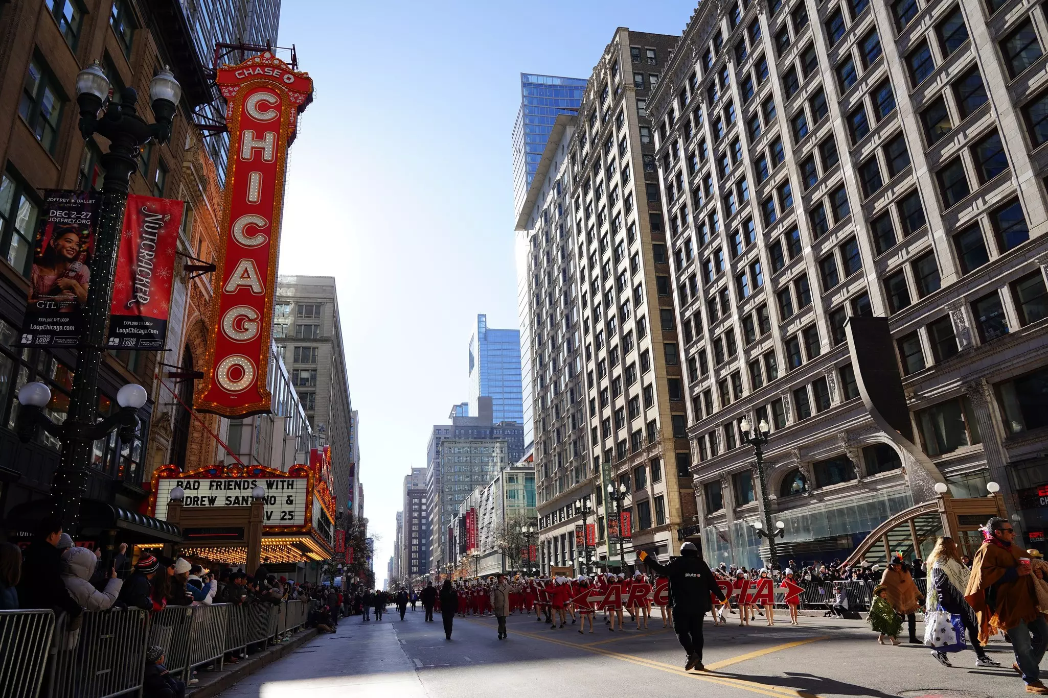 A city street with performers in the center and spectators standing behind barriers to the side.