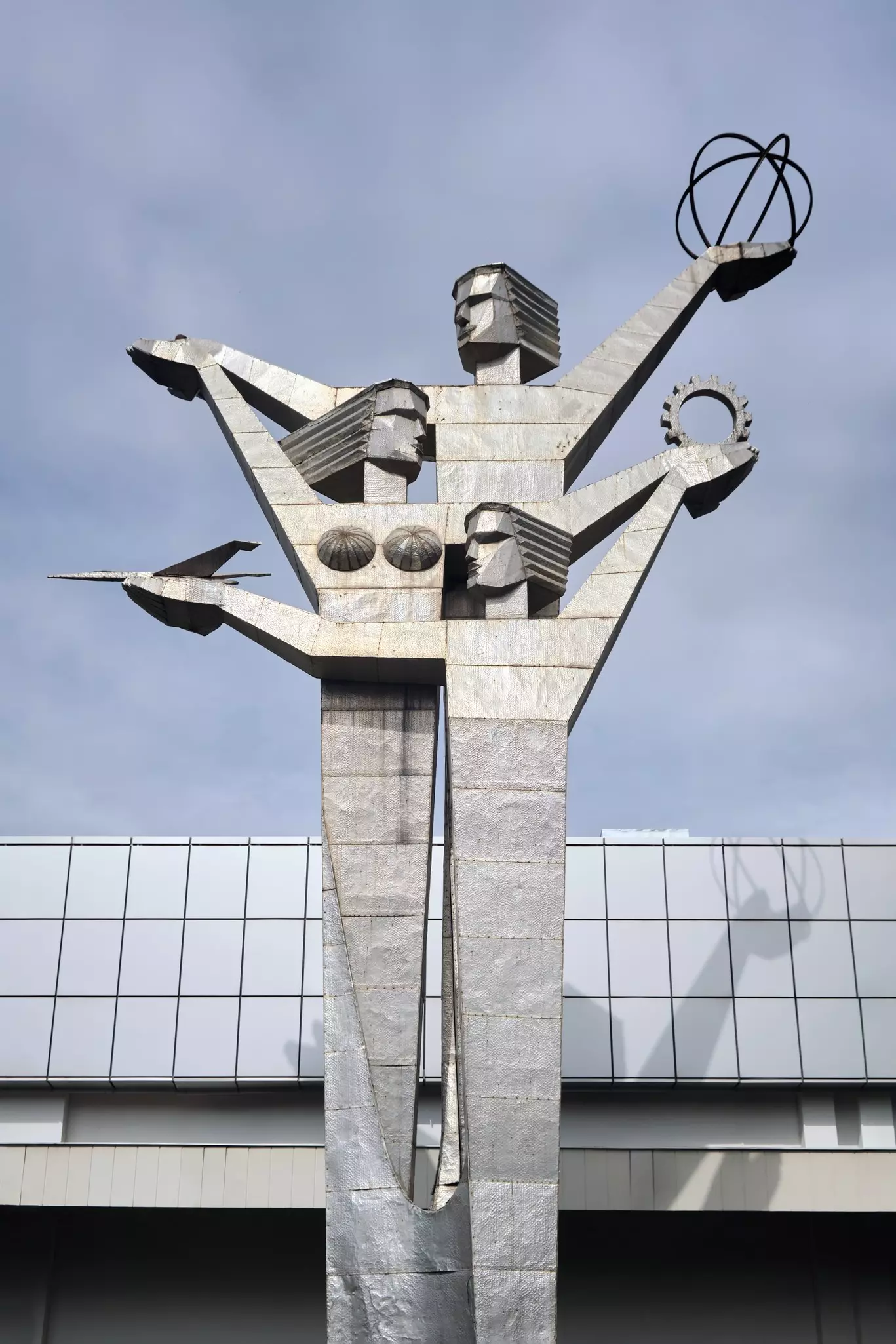 A large silver statue outside an exhibition hall, with three figures (man, woman, child) holding a globe, cog and plane high in the air.