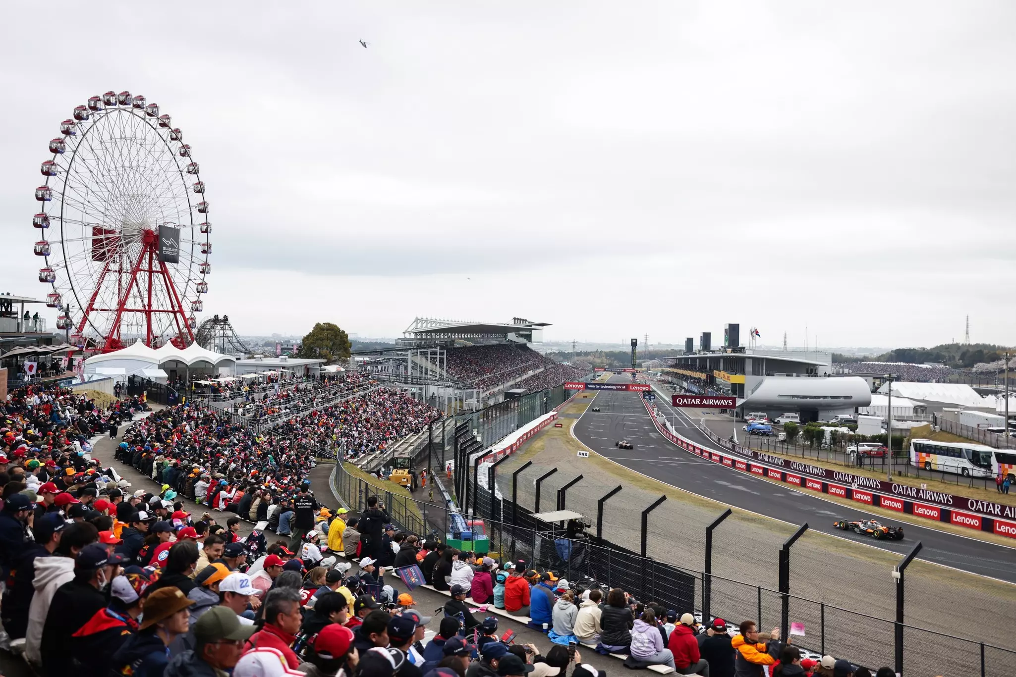 Spectators watch an auto race from the grandstands. A Ferris wheel is visible at left.