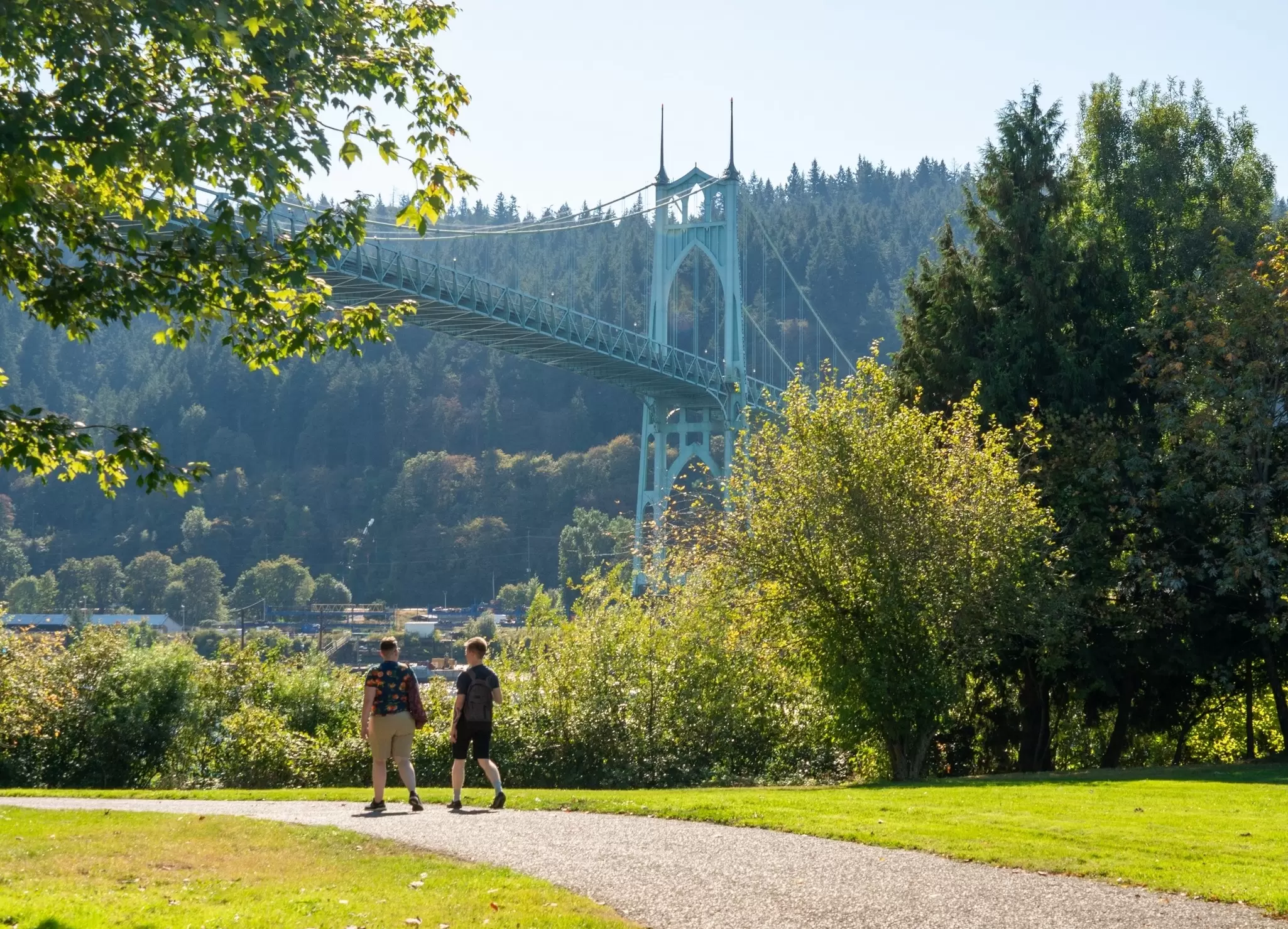 Two people walk in a park. An ornate suspension bridge is seen behind them.