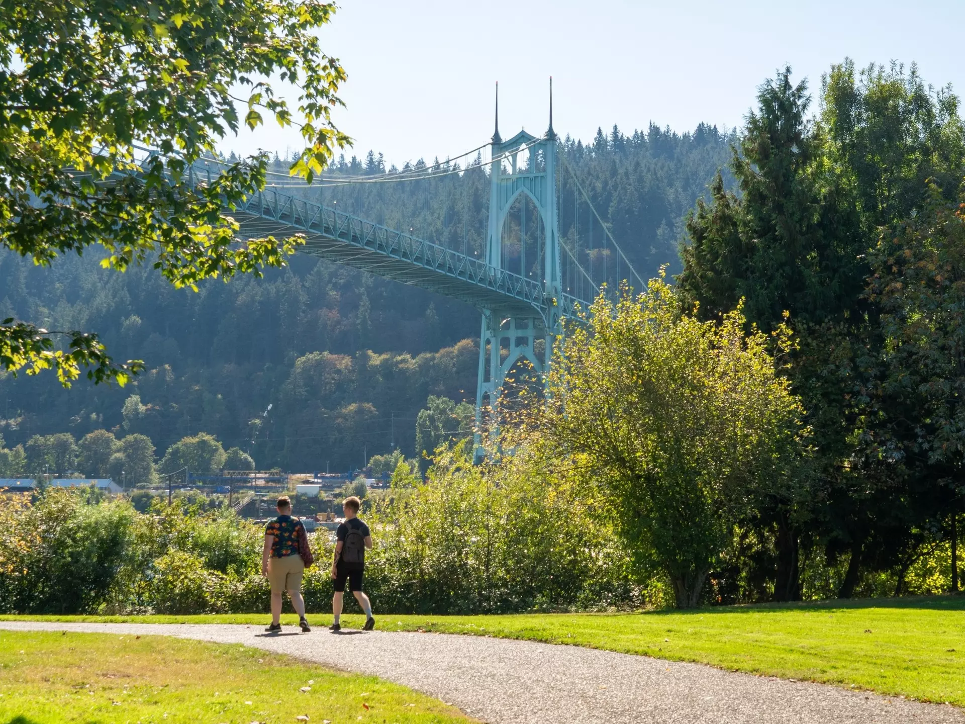 Two people walk in a park. An ornate suspension bridge is seen behind them.