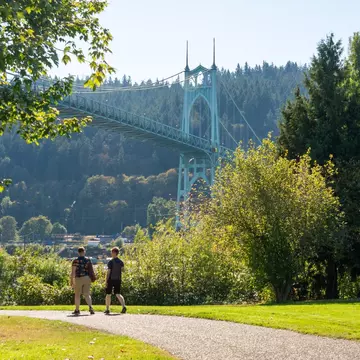 Two people walk in a park. An ornate suspension bridge is seen behind them.