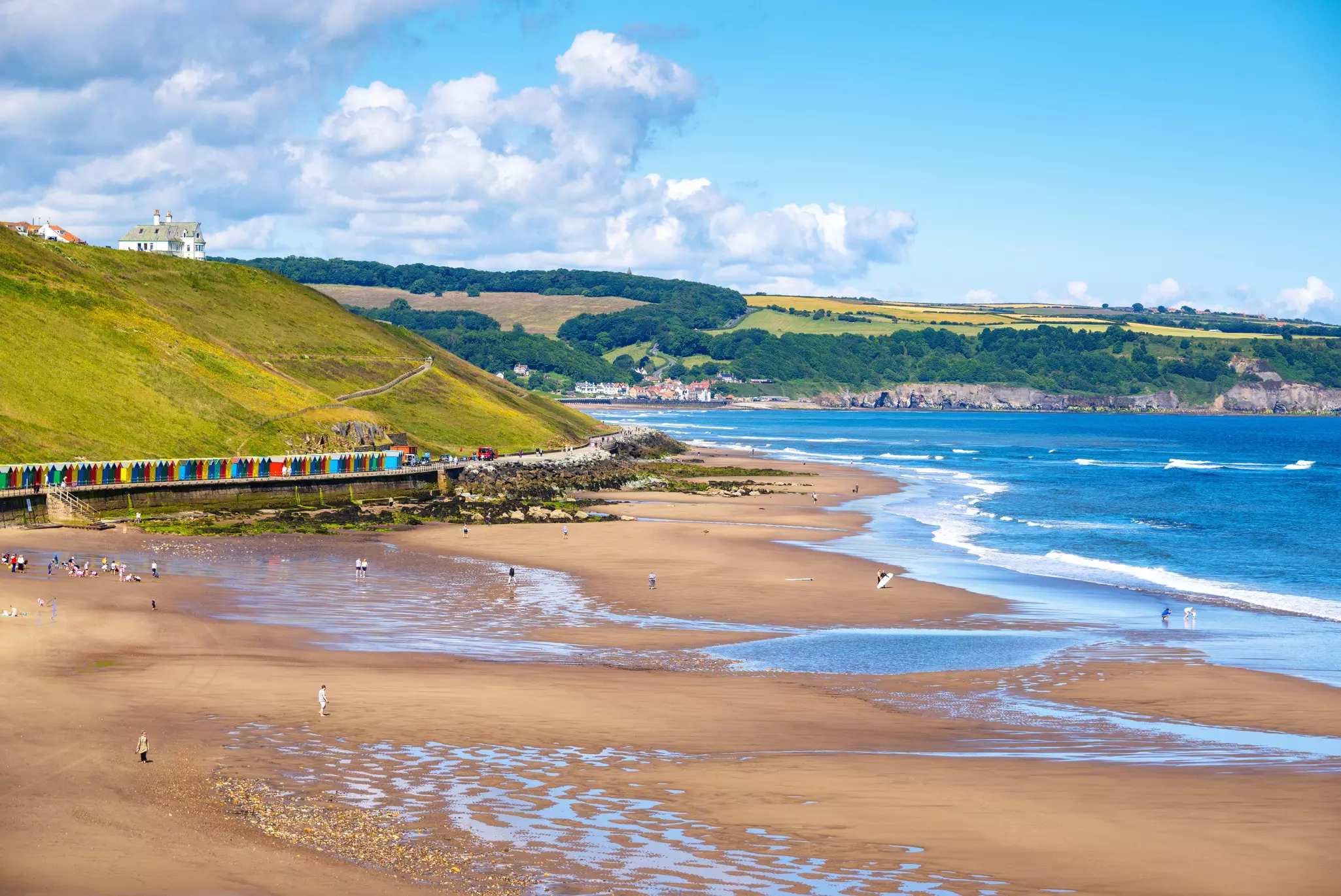 A sandy curve of sand backed by a row of colorful beach huts and grassy hills.