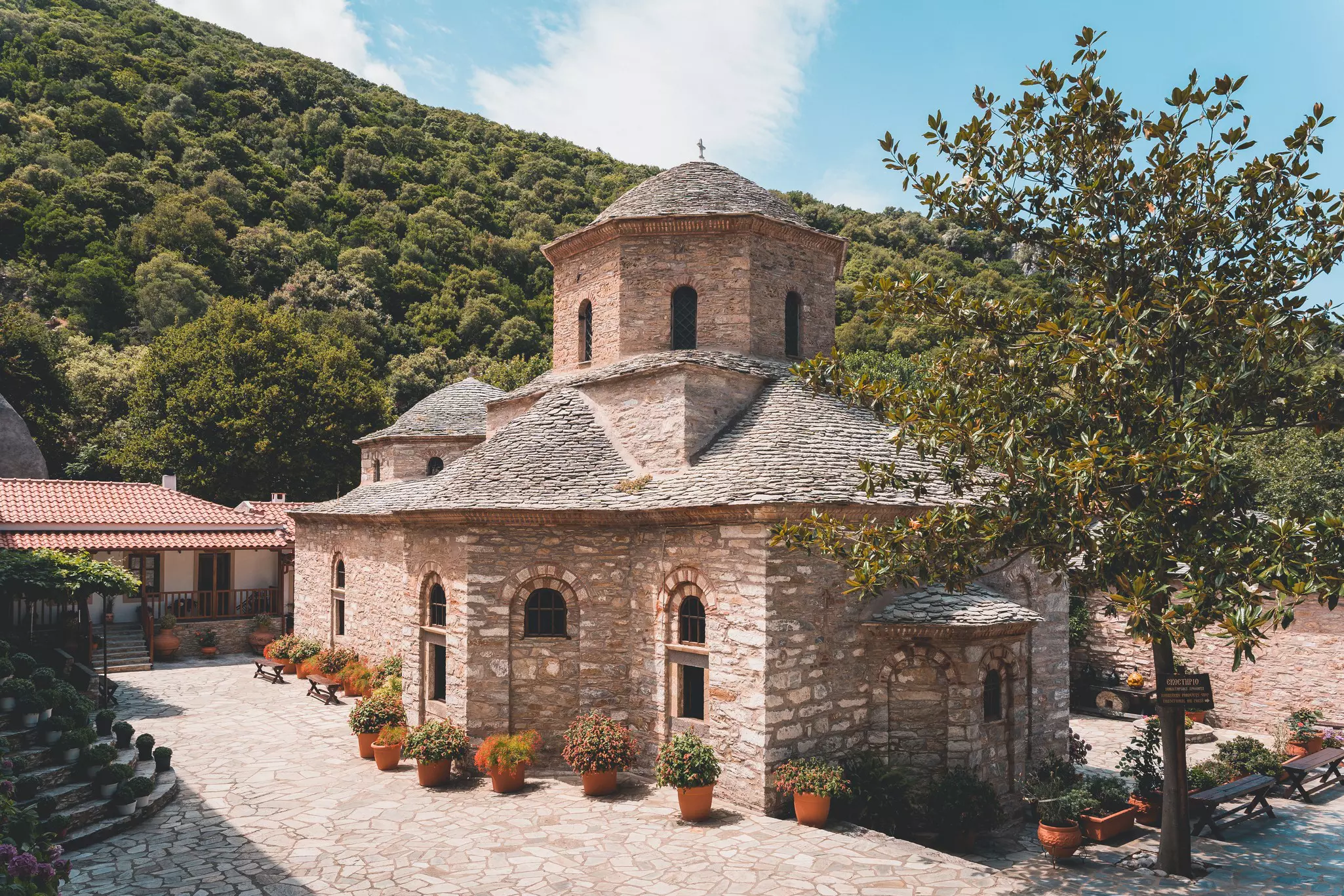 A historic stone building, part of monastery complex, is pictured against a hillside covered with trees.