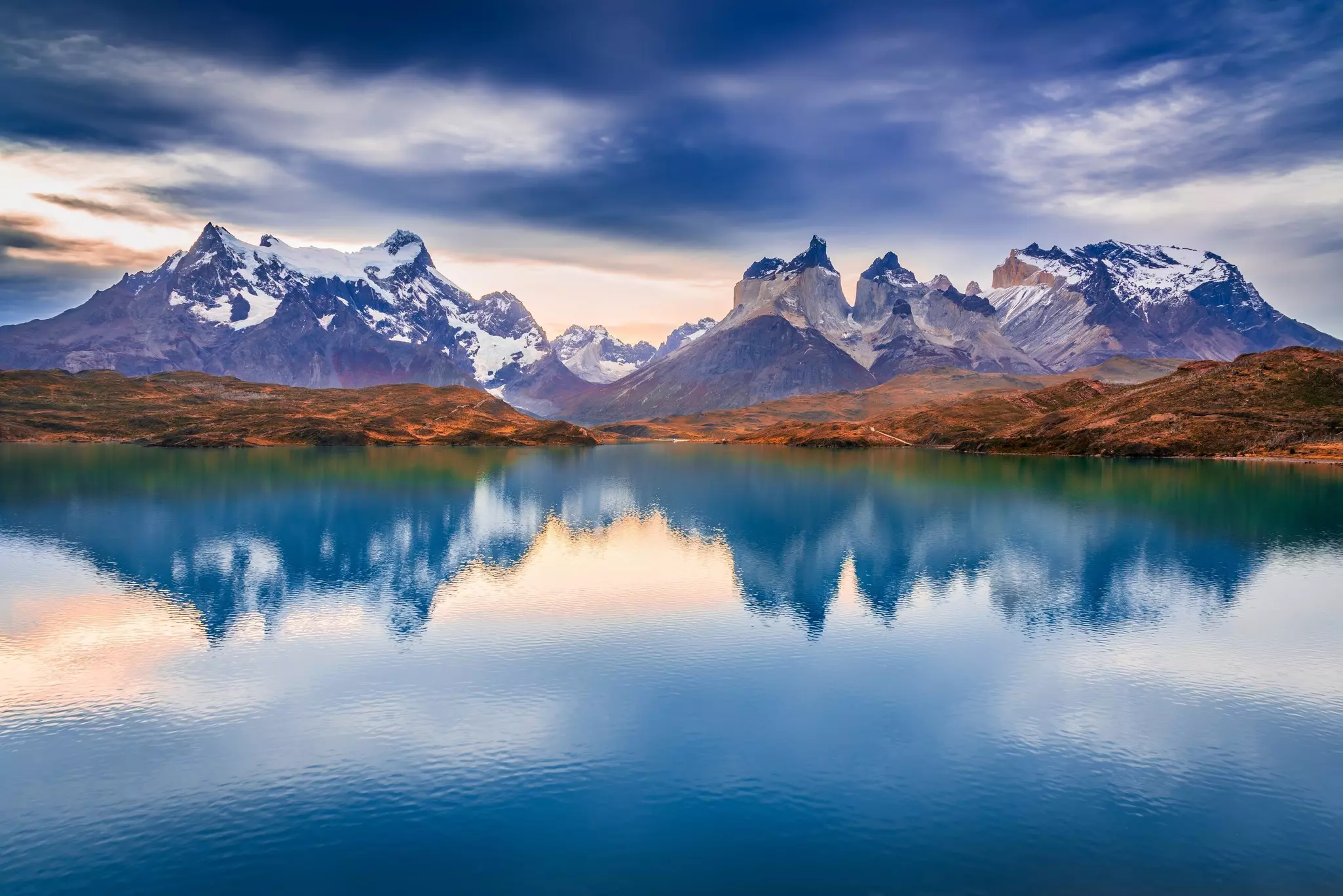 Spiky, snow-covered peaks are reflected on the surface of a lake on a cloudy day.