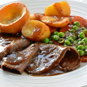 A plate of roast beef smothered in gravy with a side of Yorkshire Pudding and veggies