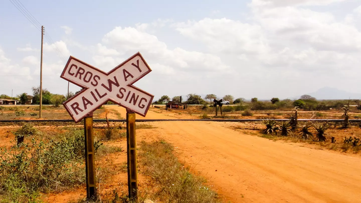 A large black-and-white cross-shaped sign reads "Railway Crossing" before a track crosses a sandy dirt road.