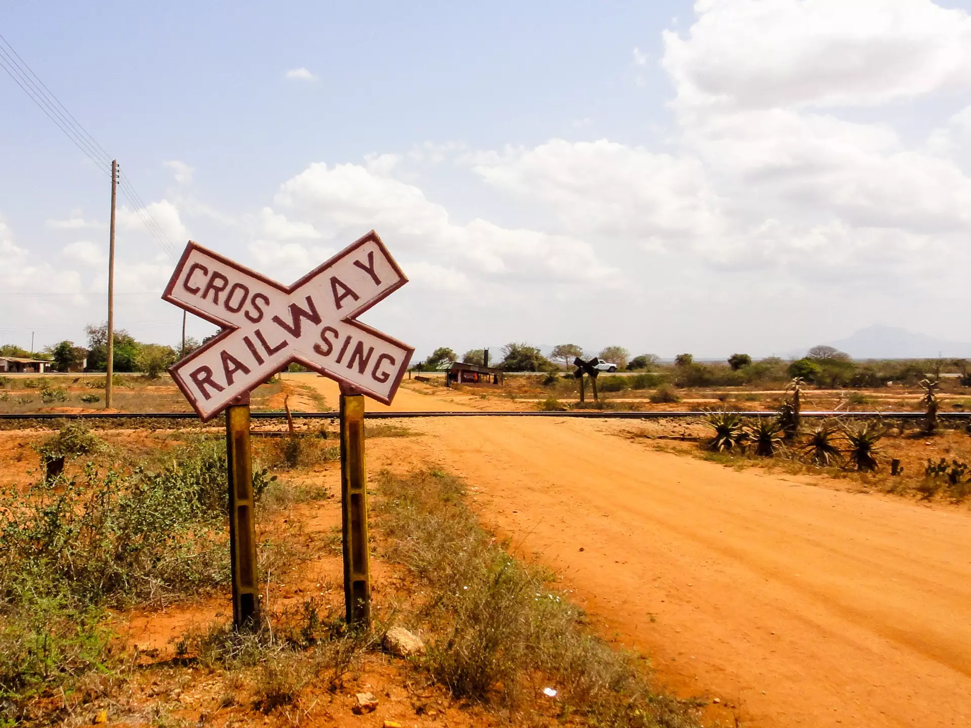 A large black-and-white cross-shaped sign reads "Railway Crossing" before a track crosses a sandy dirt road.
