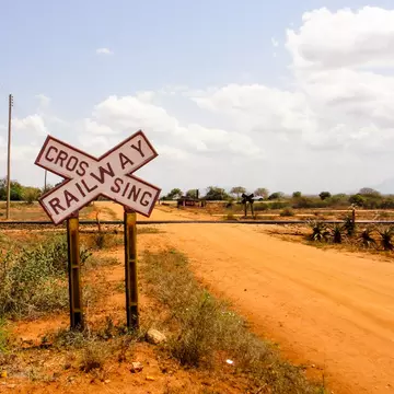 A large black-and-white cross-shaped sign reads "Railway Crossing" before a track crosses a sandy dirt road.