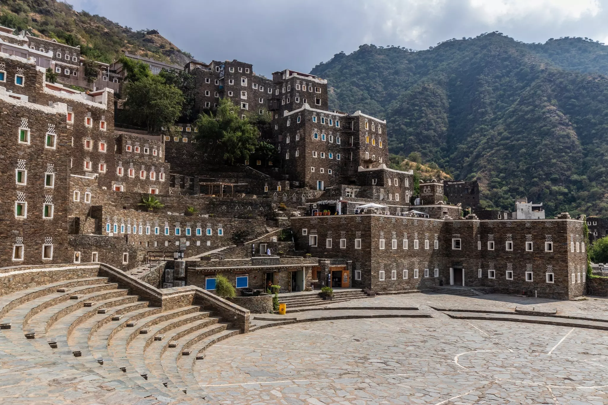Traditional stone architecture at Rijal Alma, Saudi Arabia, with forested mountains in the background.