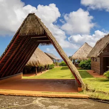 A large open-sided A-frame shelter with a steep thatched roof and red timber rafters standing on a red-earth floor, with thatched wooden huts and the sea behind it under a bright cloudy sky.