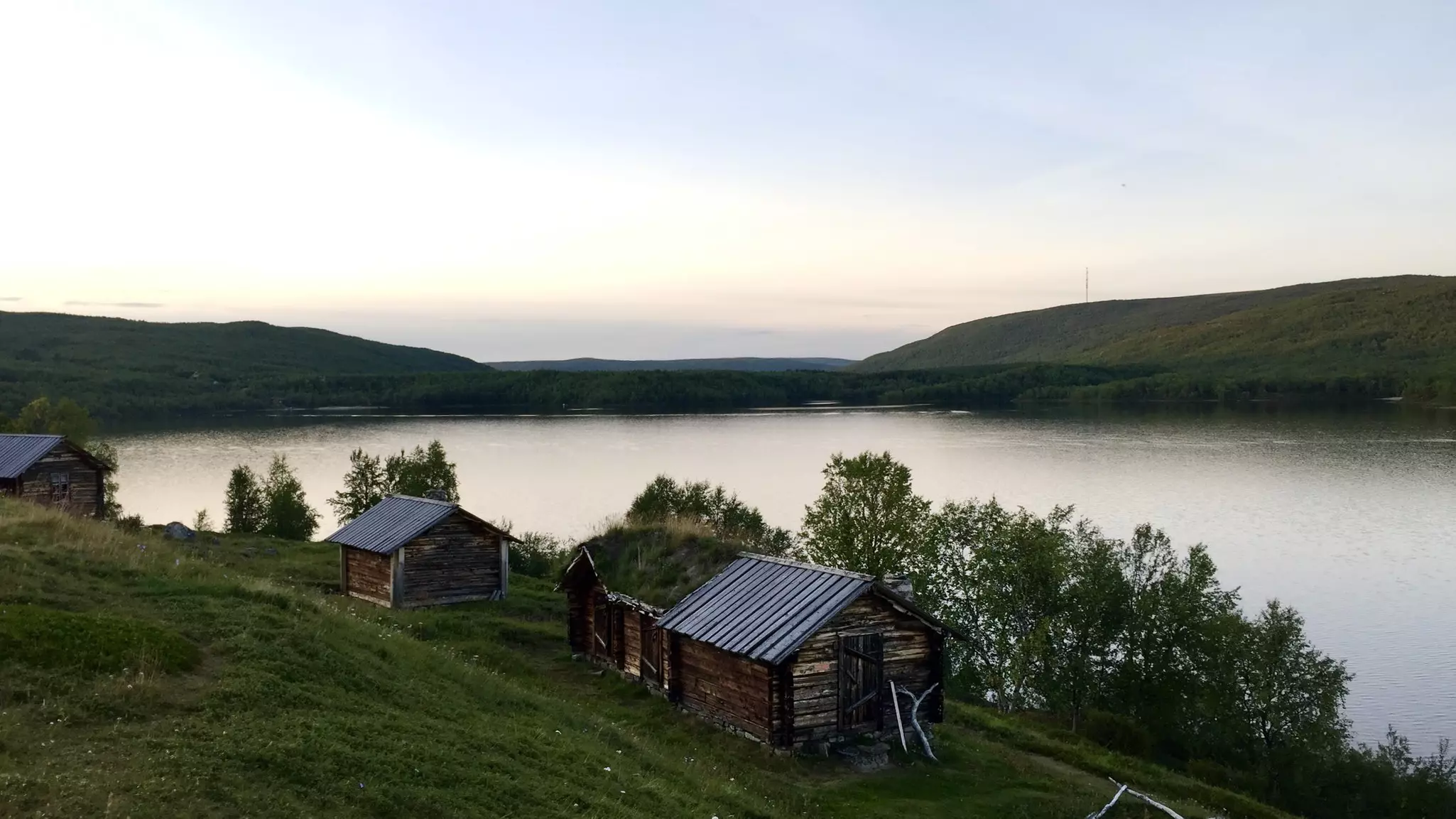 Wood buildings on a gentle green hillside by a calm lake in Finland in early evening light.
