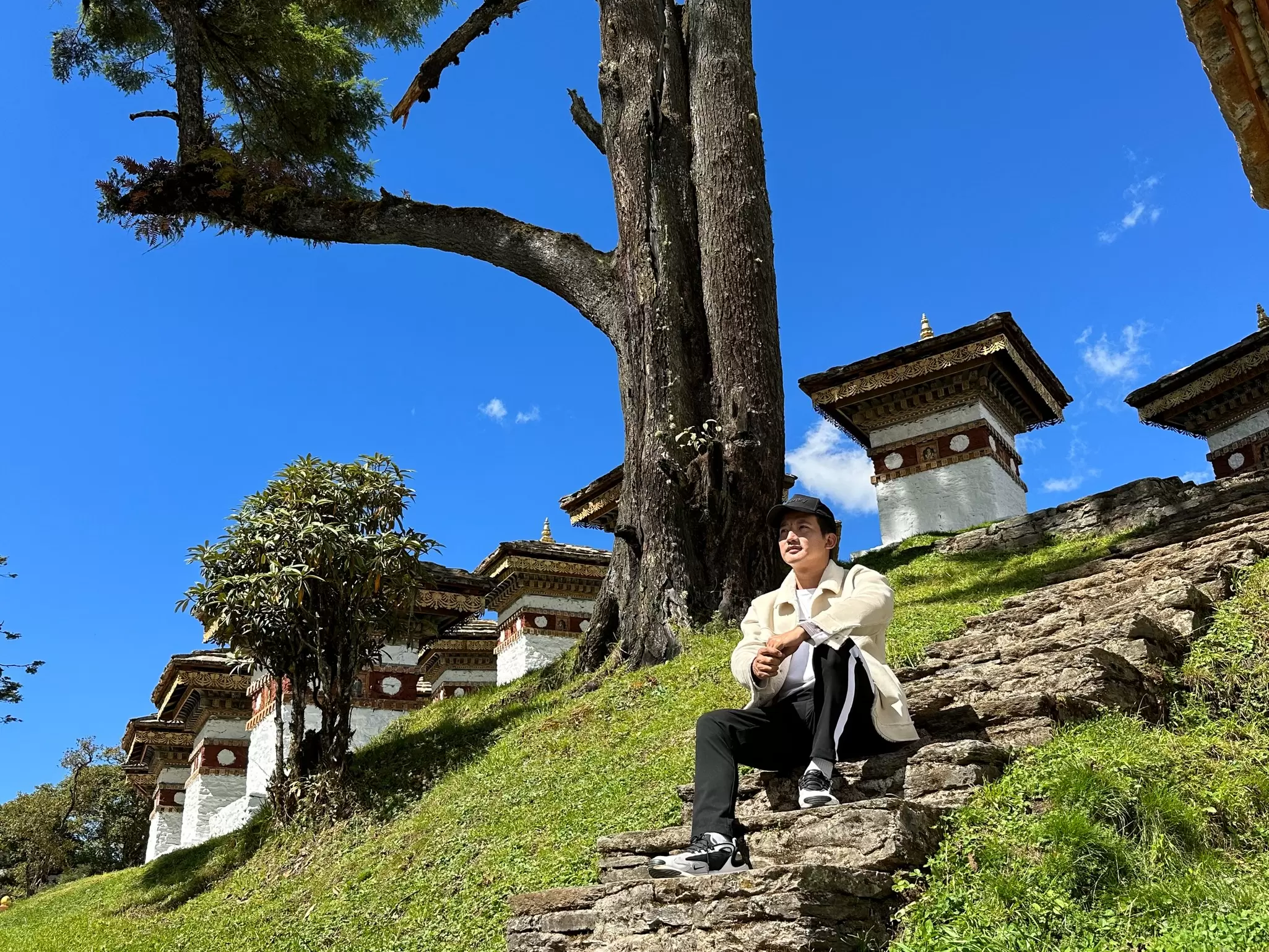 Man in a cream jacket sitting on stone steps beside whitewashed Buddhist chortens under a big tree