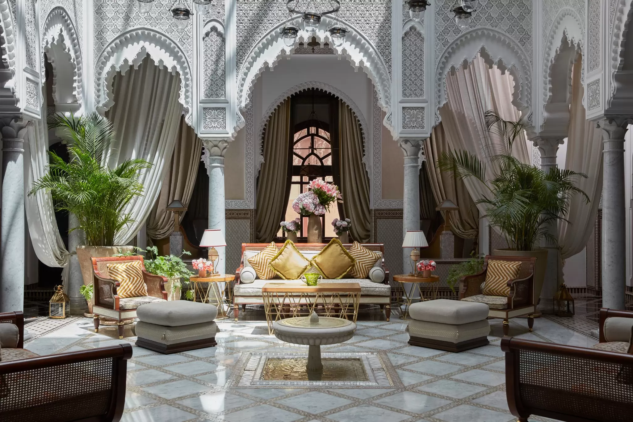 A room with large potted plants, potted flowers, ornate archways and plush furnishings, including a couch, several chairs and two ottomans.